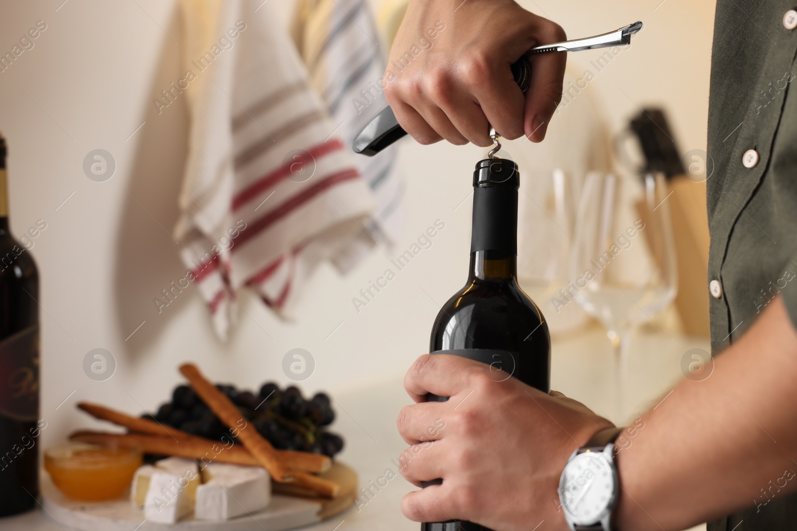 Photo of Romantic dinner. Man opening wine bottle with corkscrew in kitchen, closeup