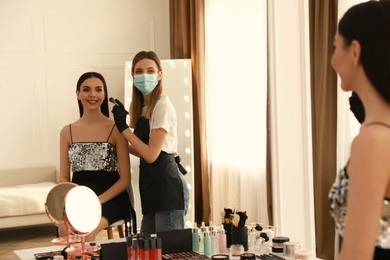 Photo of Makeup artist in protective mask and gloves working with woman indoors. Preventive measures during COVID-19 pandemic