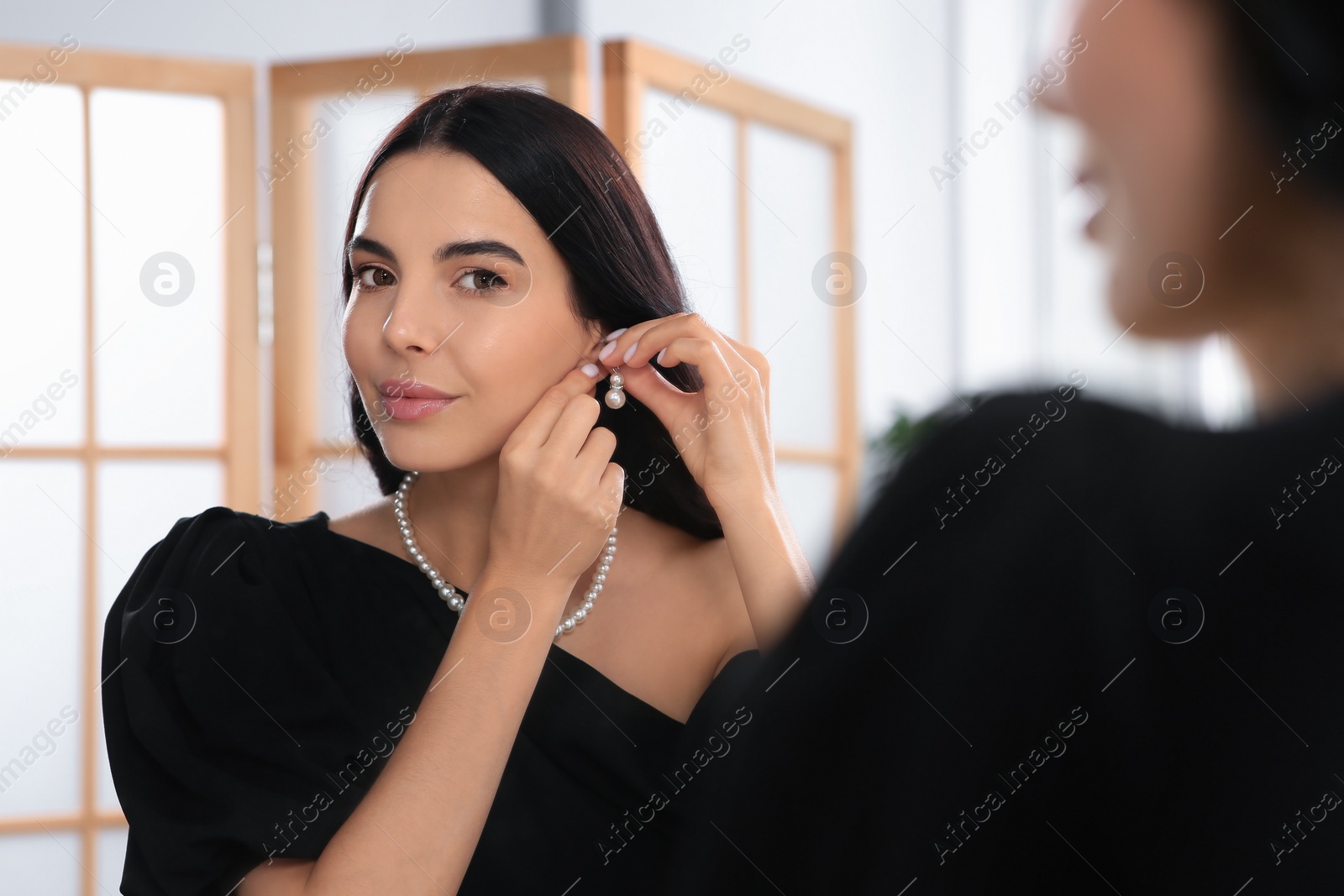 Photo of Young woman trying on elegant pearl earring near mirror indoors