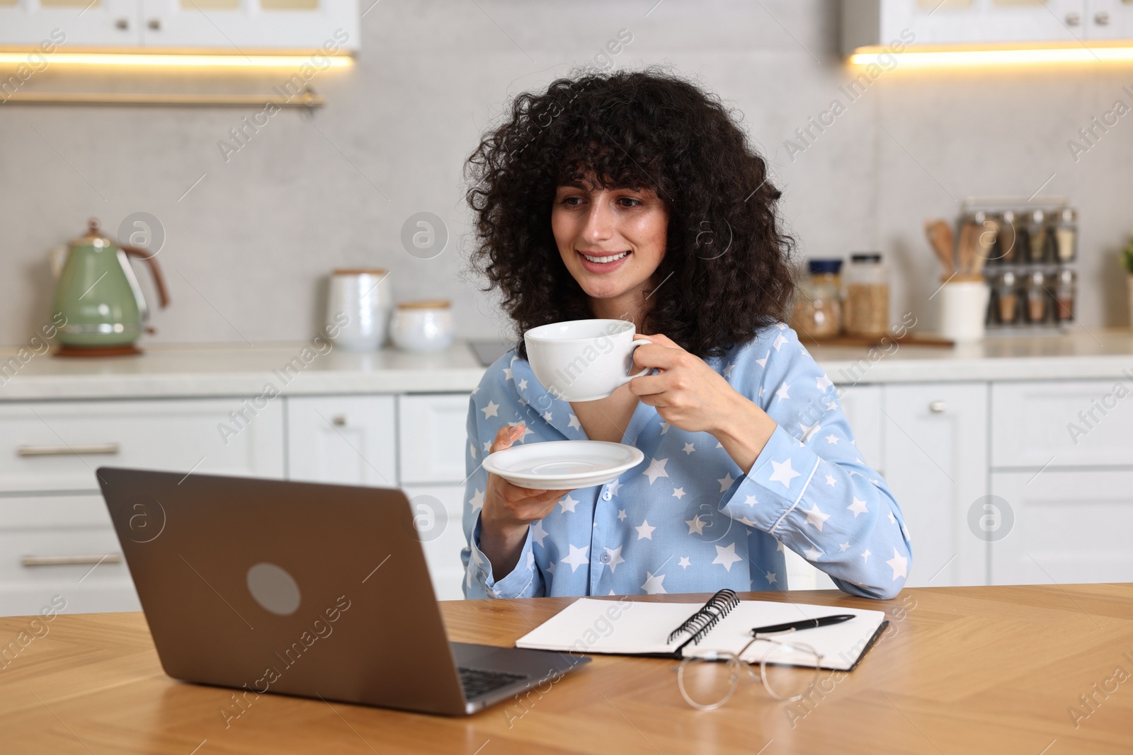 Photo of Beautiful young woman in stylish pyjama with cup of drink using laptop at wooden table in kitchen