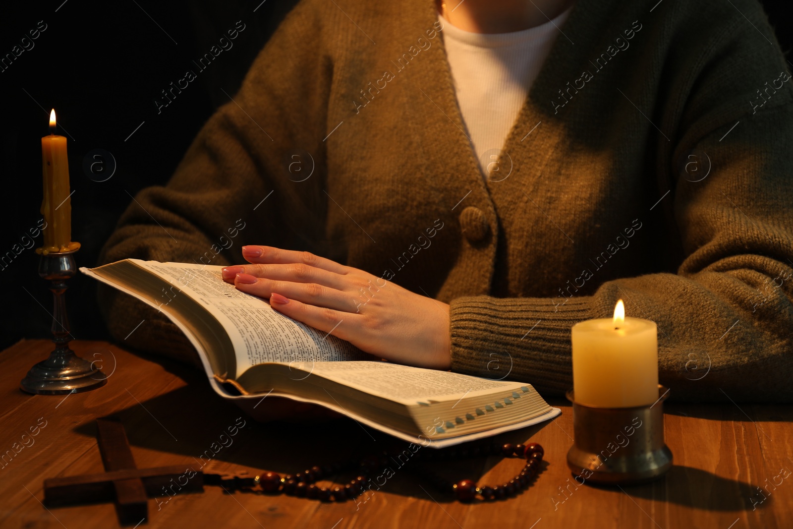 Photo of Woman reading Bible at table with burning candles, closeup