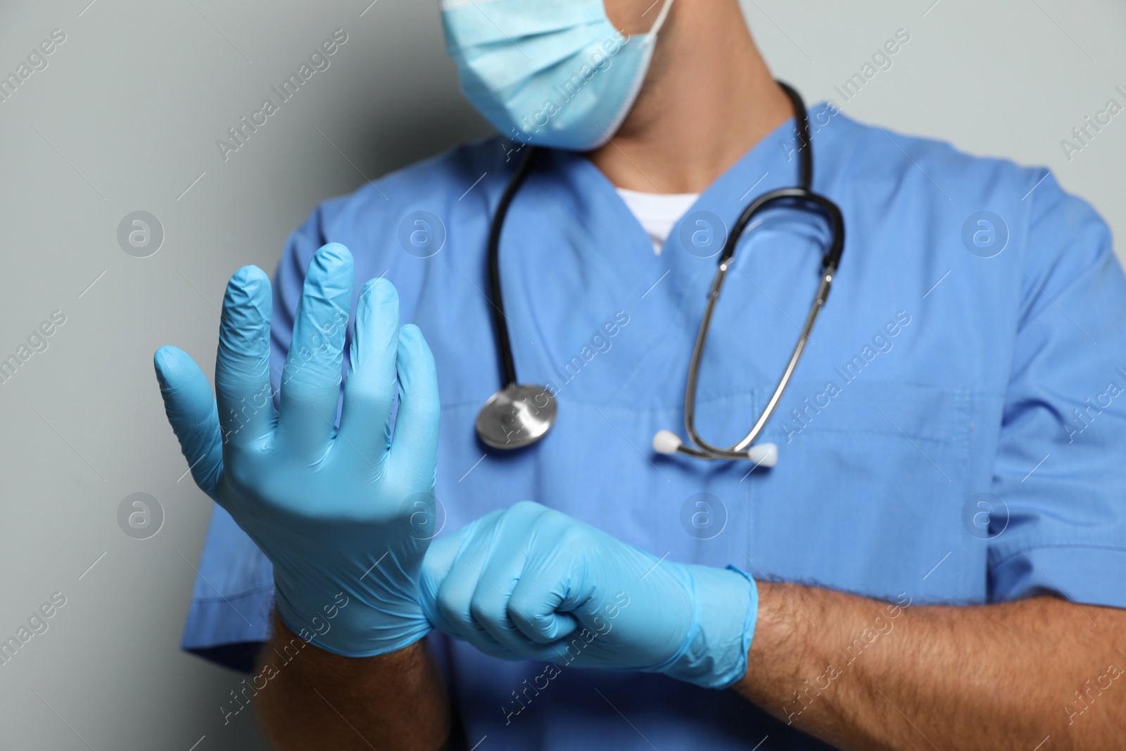 Photo of Doctor in protective mask putting on medical gloves against light grey background, closeup