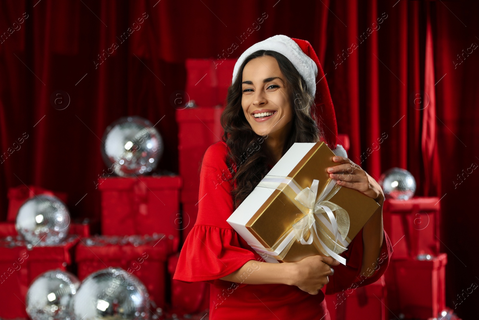 Photo of Beautiful woman in Christmas costume with gift near pile of presents indoors