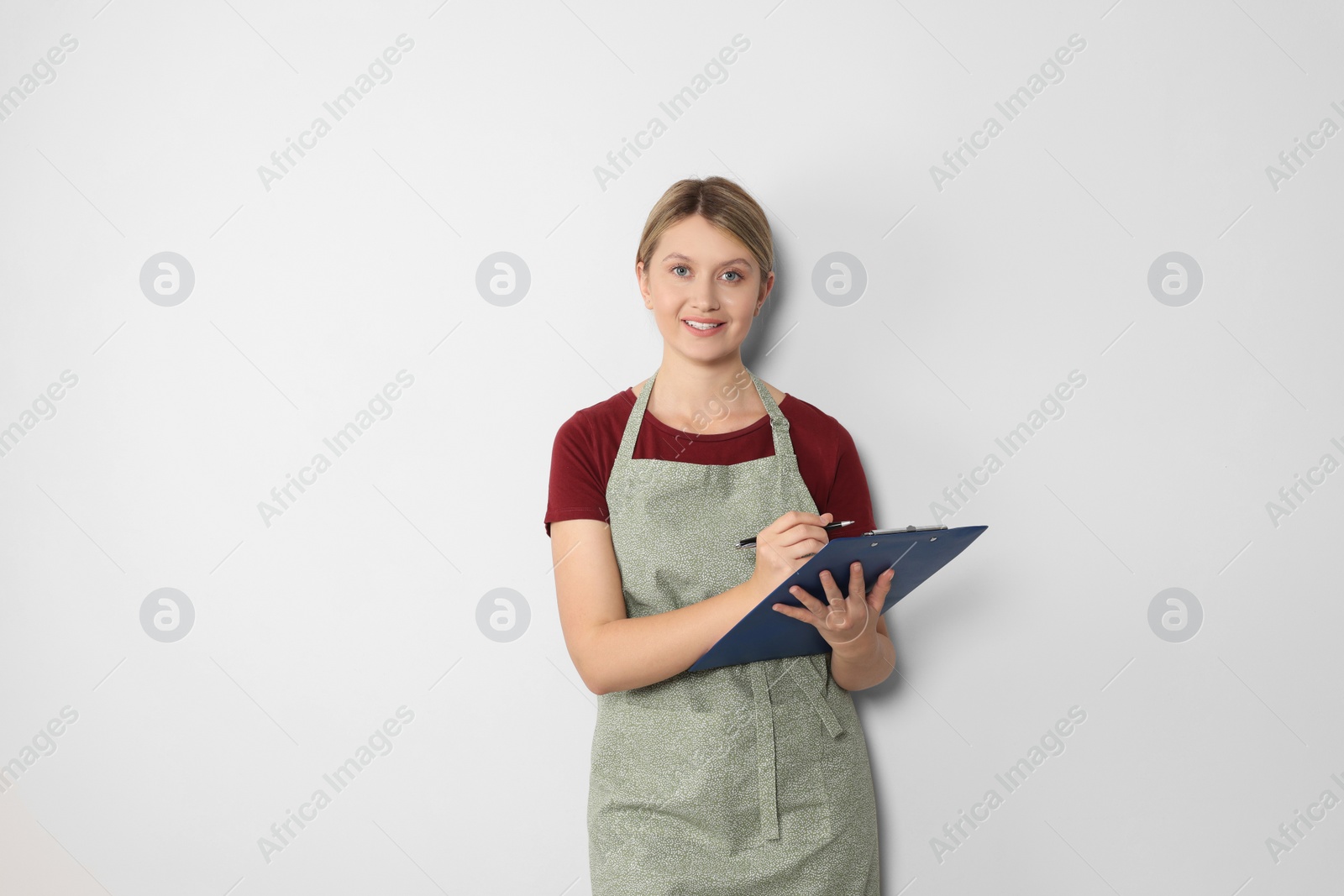 Photo of Beautiful young woman in clean apron with clipboard on light grey background