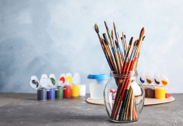 Photo of Glass jar with brushes and different paints on table against color background. Space for text