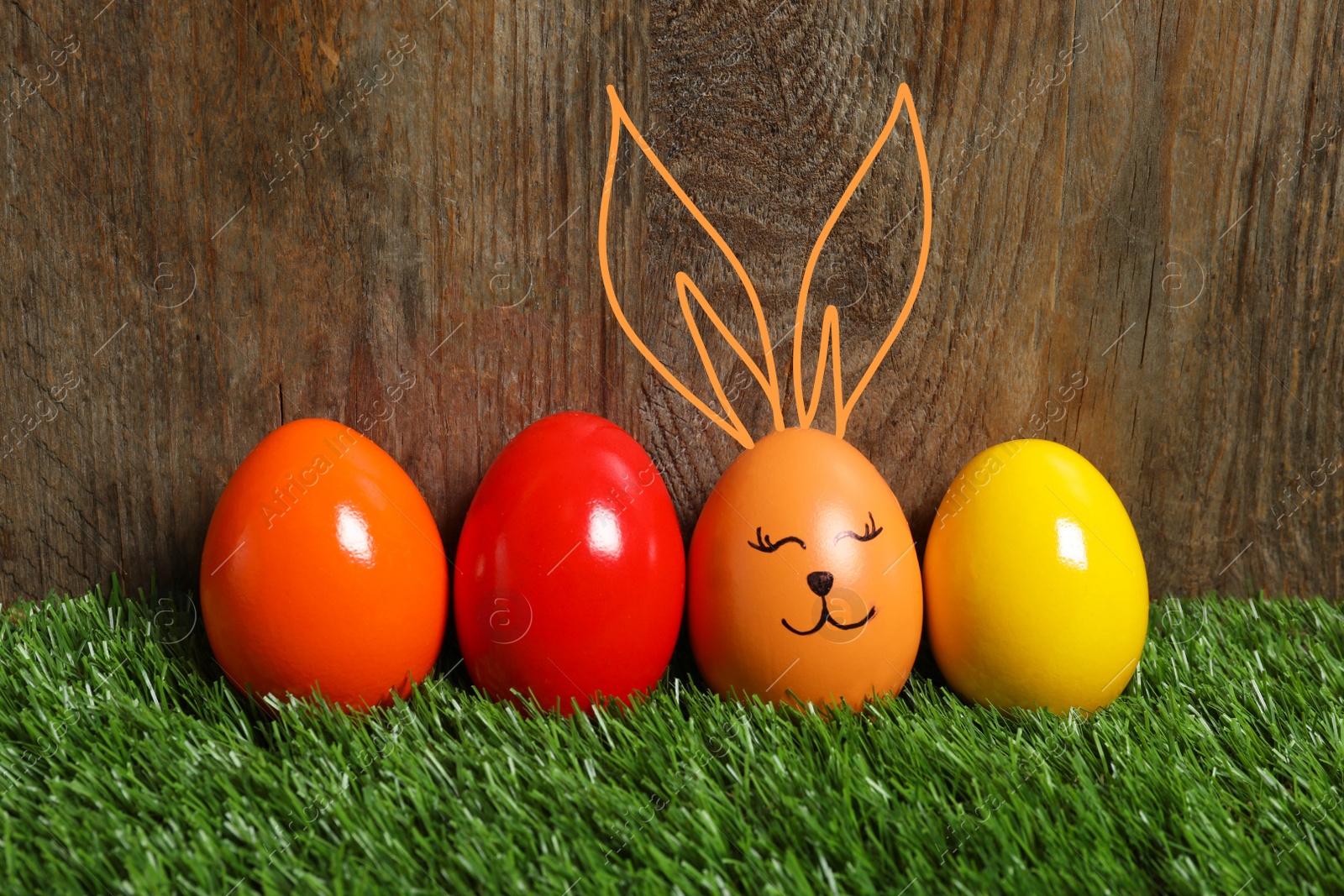 Image of One egg with drawn face and ears as Easter bunny among others on green grass against wooden background