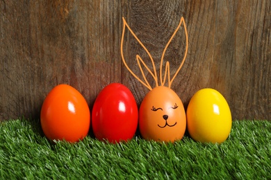 Image of One egg with drawn face and ears as Easter bunny among others on green grass against wooden background
