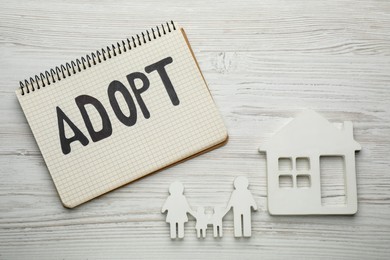 Photo of Family figure, house and notebook with word Adopt on white wooden table, flat lay