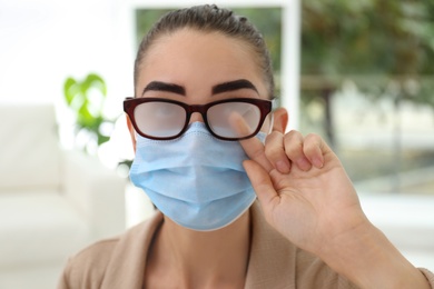 Photo of Woman wiping foggy glasses caused by wearing medical mask indoors, closeup