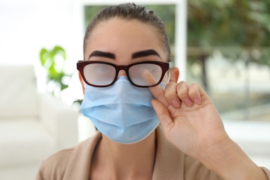 Woman wiping foggy glasses caused by wearing medical mask indoors, closeup