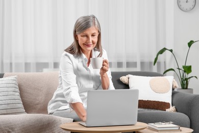 Photo of Beautiful senior woman with cup of drink using laptop at home