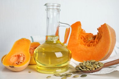 Photo of Glass jug of oil with cut pumpkin and seeds on white wooden table