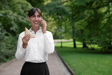 Photo of Lunch time. Happy businesswoman with sandwich talking on smartphone in park, space for text