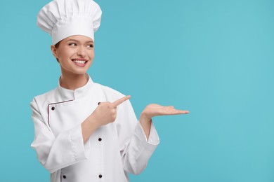 Photo of Happy woman chef in uniform pointing at something on light blue background, space for text