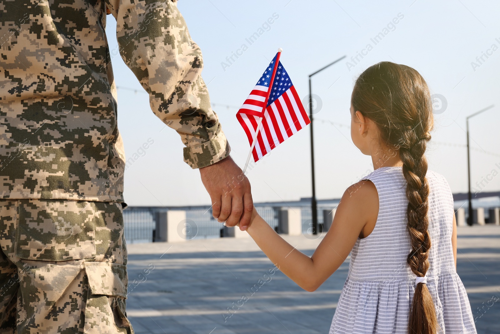 Photo of Soldier and his little daughter with American flag holding hands outdoors, back view. Veterans Day in USA