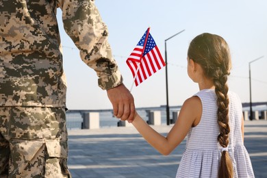 Photo of Soldier and his little daughter with American flag holding hands outdoors, back view. Veterans Day in USA