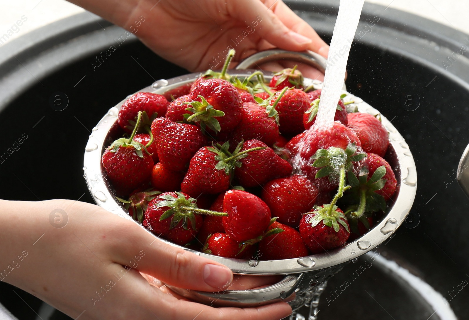 Photo of Woman washing fresh strawberries under tap water in metal colander above sink, closeup