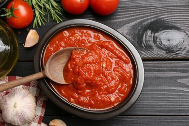 Photo of Homemade tomato sauce in bowl, spoon and fresh ingredients on black wooden table, flat lay