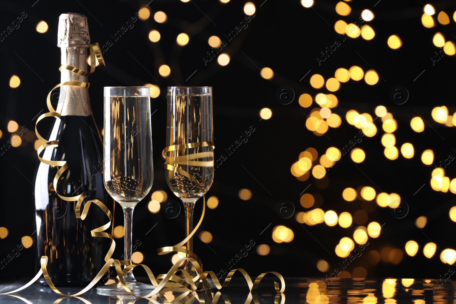 Photo of Glasses and bottle of champagne with serpentine streamers against blurred lights on black background. Space for text