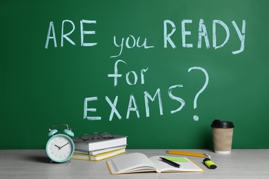 Photo of Different stationery and alarm clock on wooden table near chalkboard with phrase Are You Ready For Exams