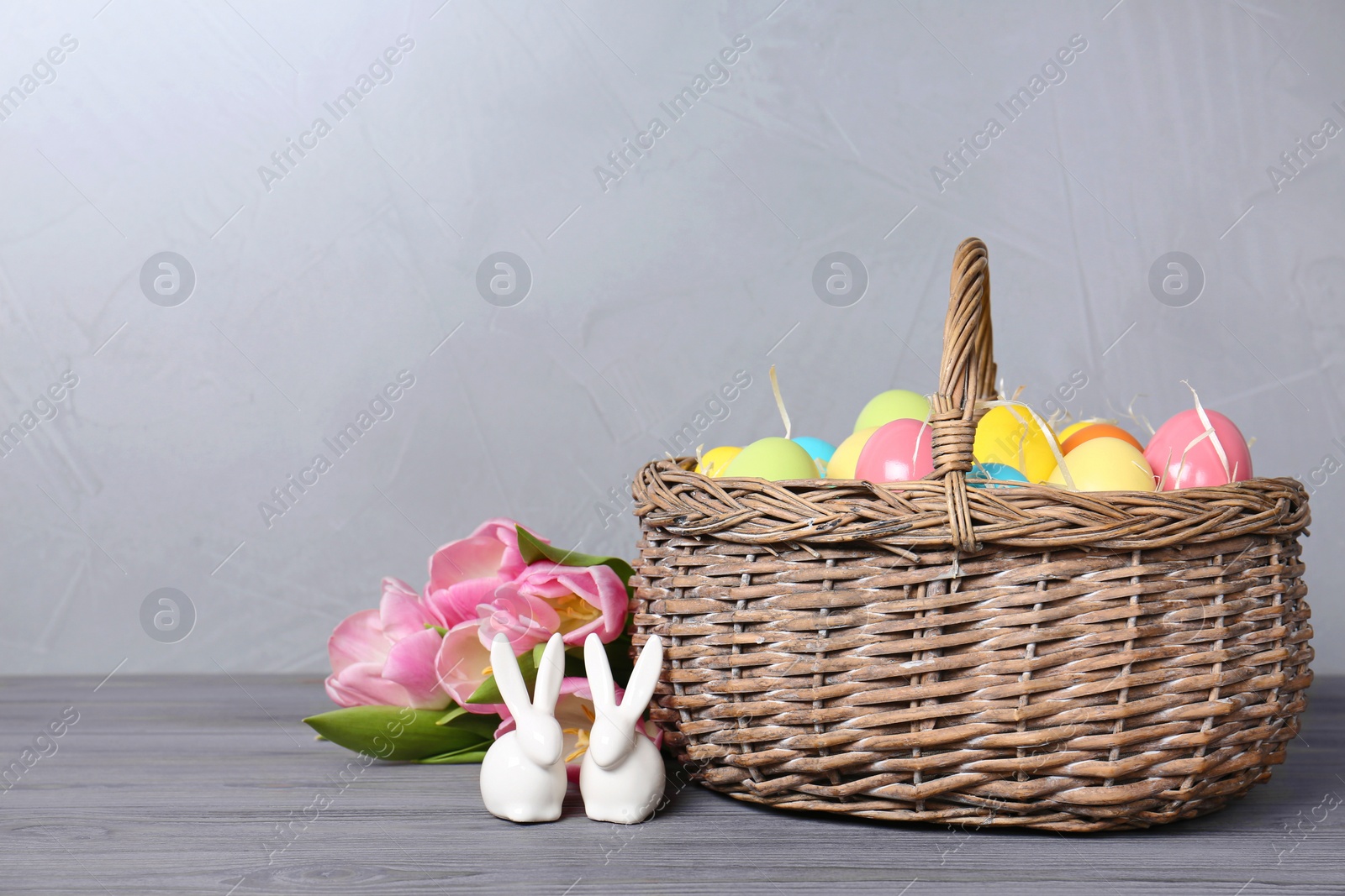 Photo of Ceramic bunnies near wicker basket with Easter eggs and spring tulips on wooden table against light background, space for text