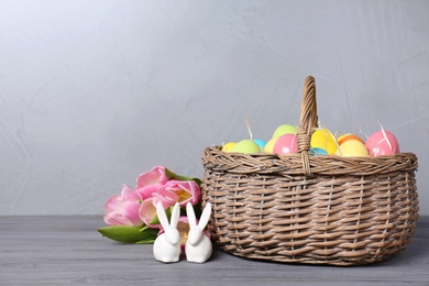 Ceramic bunnies near wicker basket with Easter eggs and spring tulips on wooden table against light background, space for text