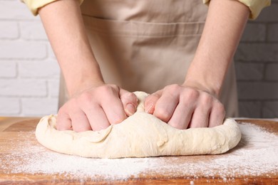 Photo of Woman kneading dough at wooden table near white brick wall, closeup