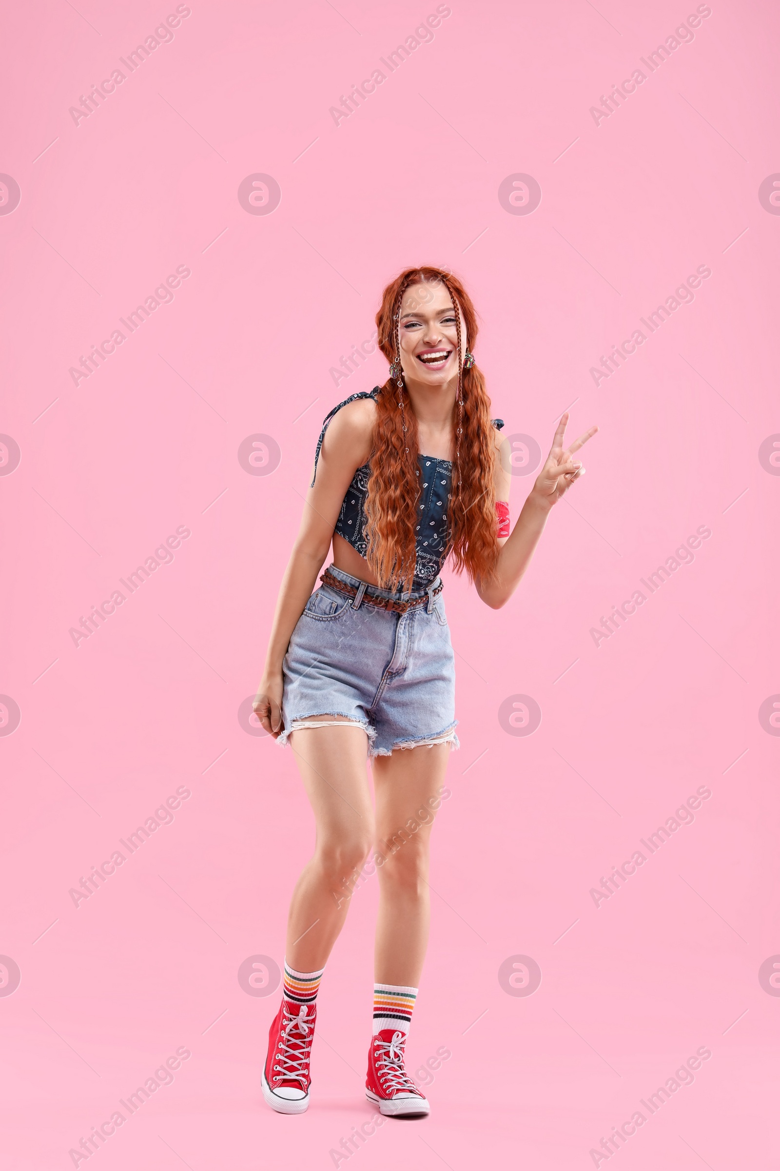 Photo of Stylish young hippie woman showing V-sign on pink background