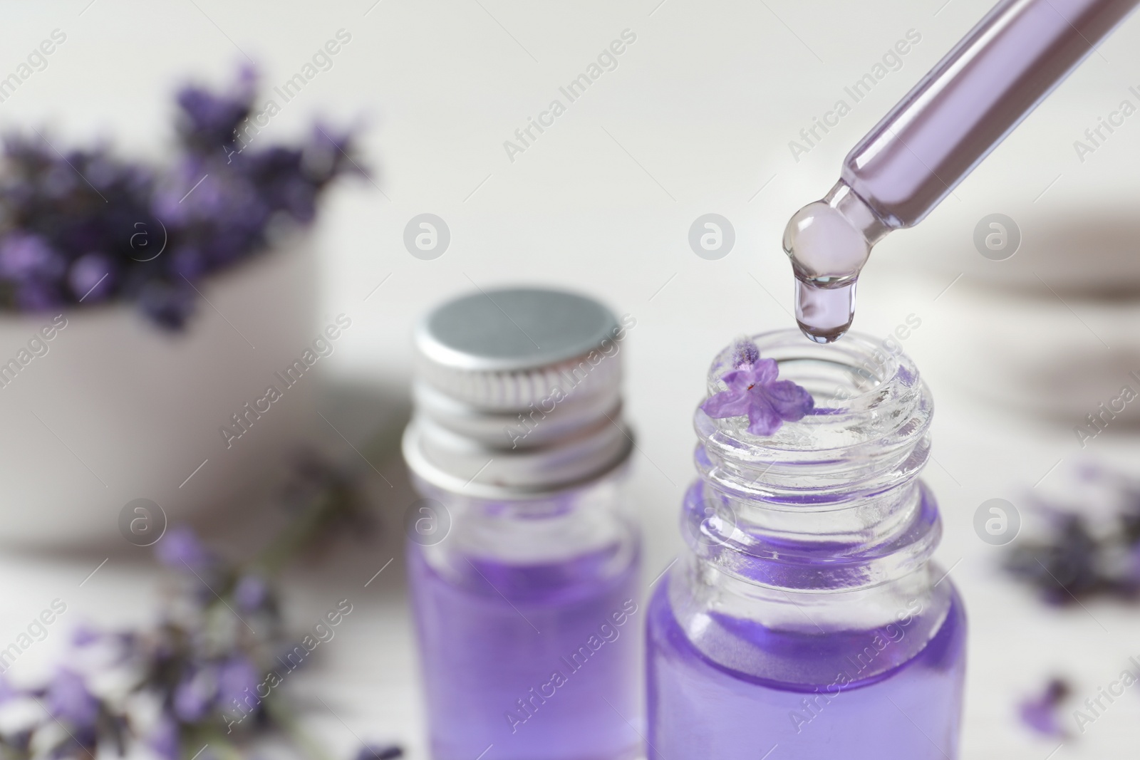 Photo of Dripping natural cosmetic oil into glass bottle with lavender flower on blurred background, closeup. Space for text