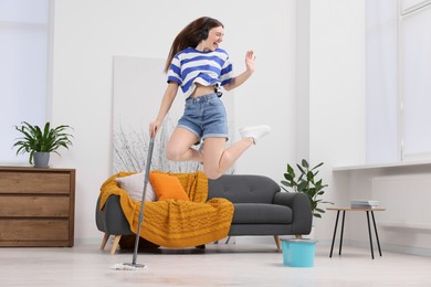 Photo of Enjoying cleaning. Happy woman in headphones jumping with mop at home