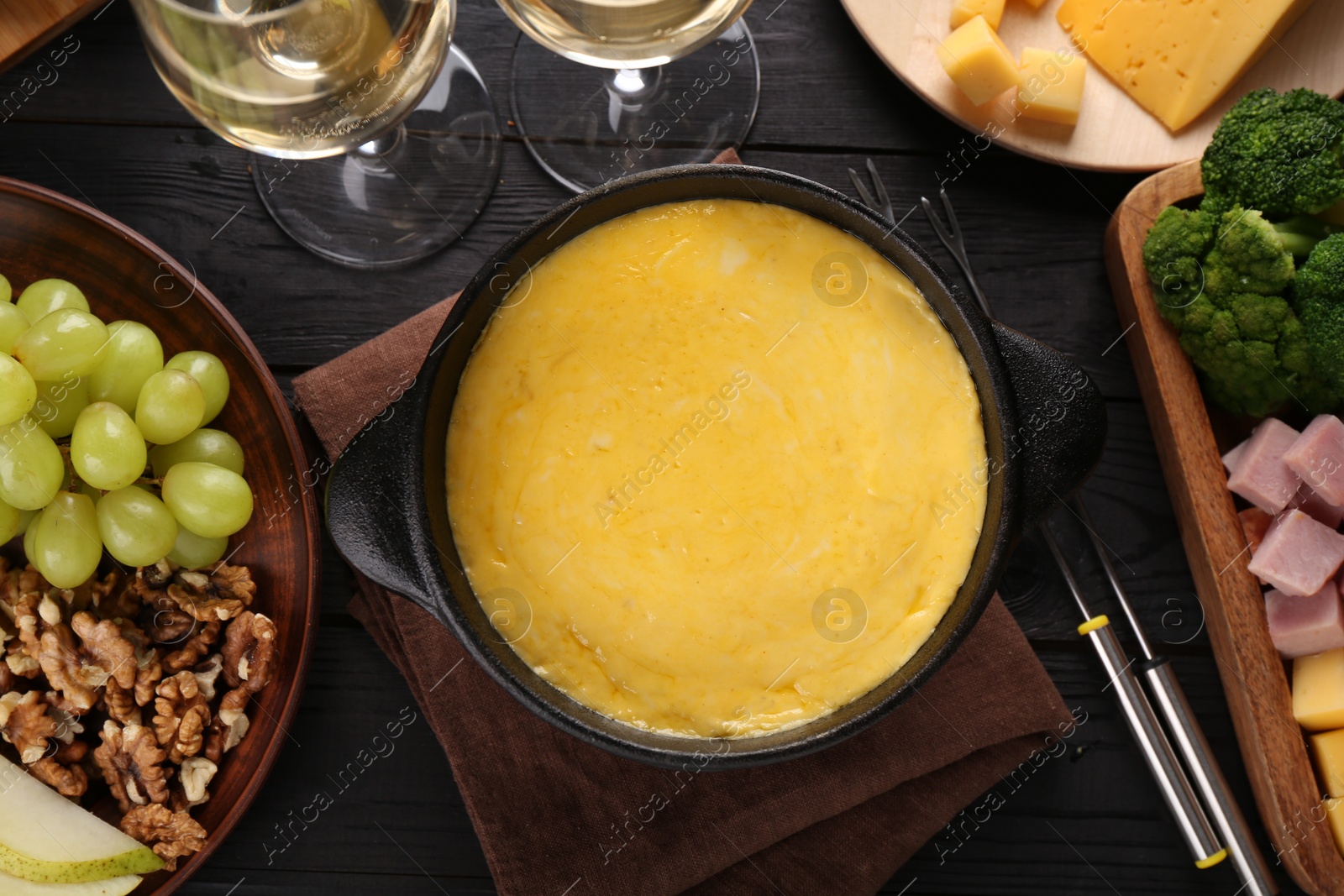 Photo of Fondue pot with melted cheese, glasses of wine and different products on black wooden table, flat lay