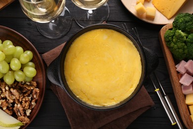 Photo of Fondue pot with melted cheese, glasses of wine and different products on black wooden table, flat lay