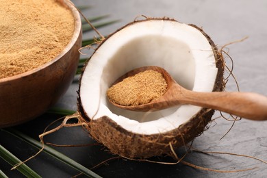 Photo of Spoon with coconut sugar, fruit, bowl and palm leaves on dark textured table, closeup