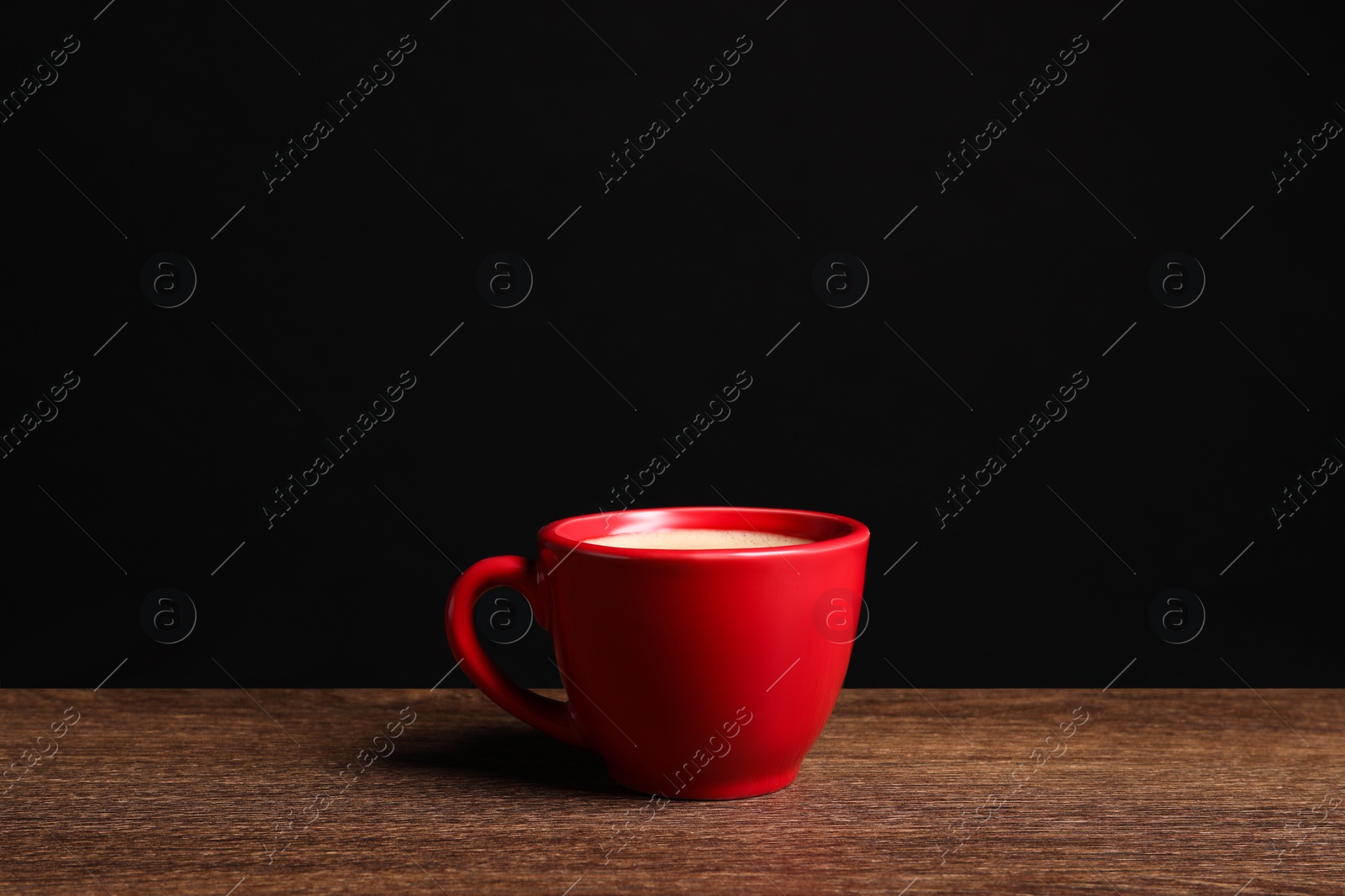 Photo of Red ceramic cup with hot aromatic coffee on wooden table against black background