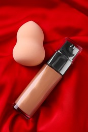 Photo of Makeup sponge and skin foundation on red cloth, flat lay