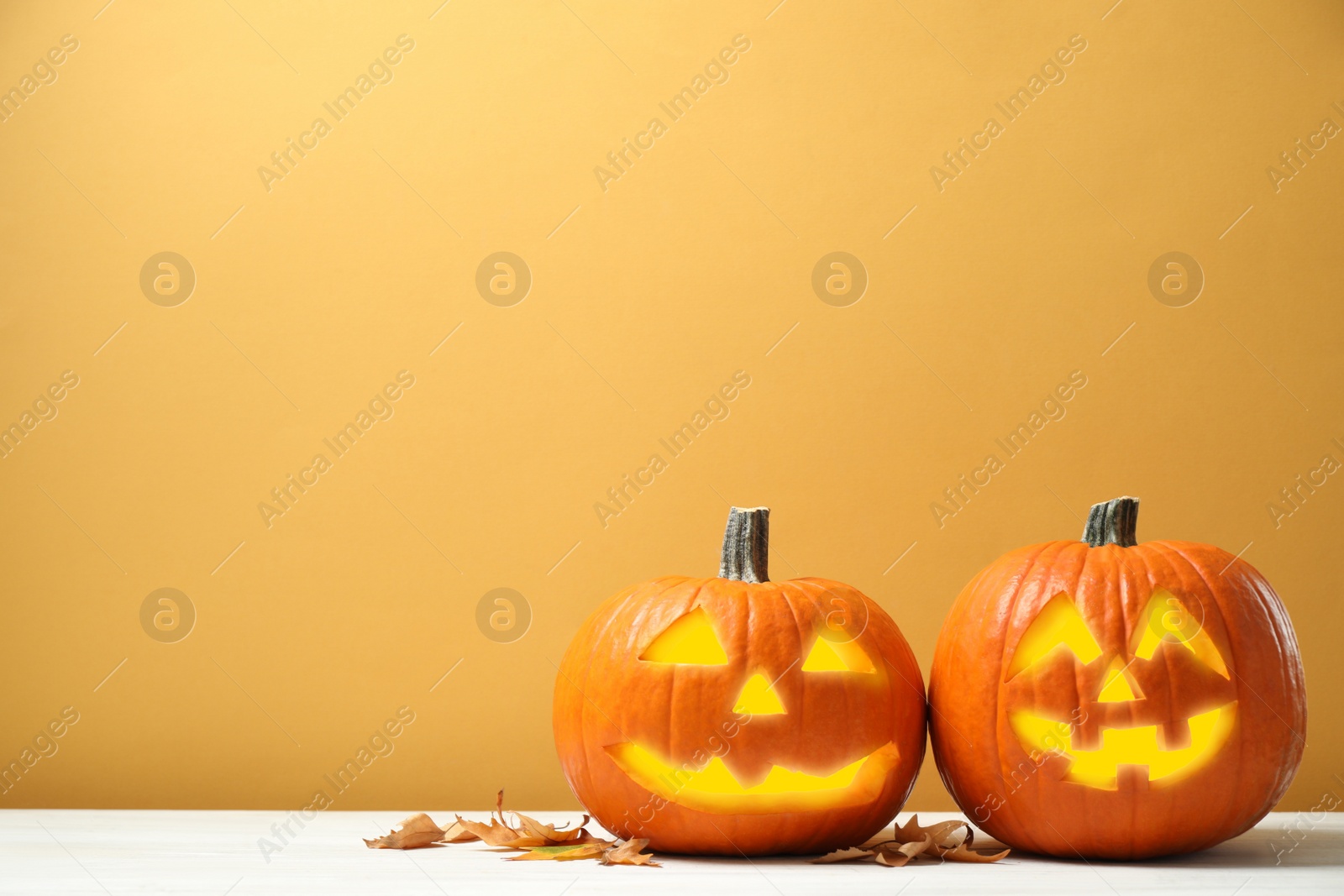 Photo of Scary jack o'lantern pumpkins and fallen leaves on white wooden table against yellow background, space for text. Halloween decor