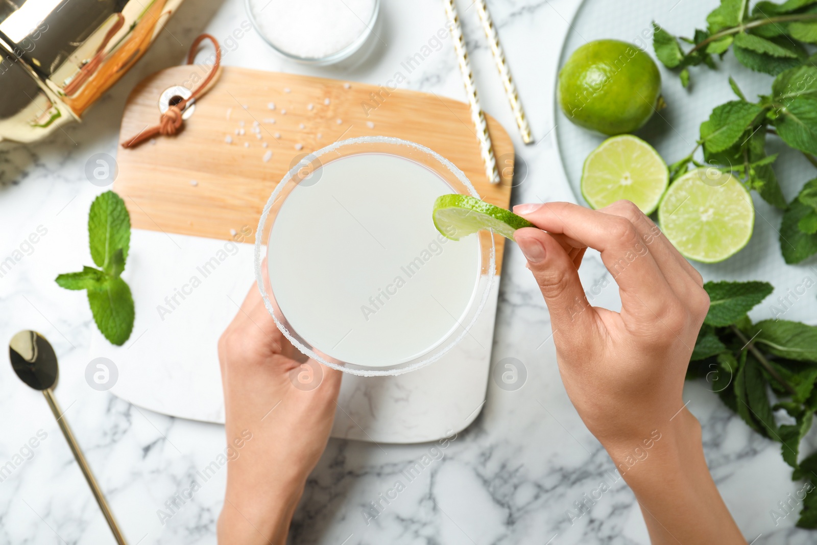 Photo of Woman decorating freshly made cocktail with lime at white marble table, top view