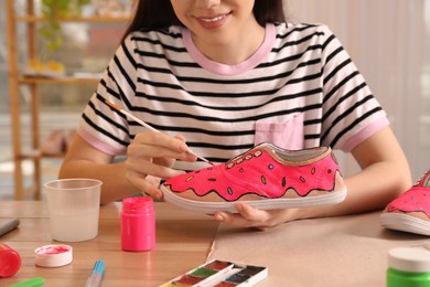 Photo of Woman painting on sneaker at wooden table indoors, closeup. Customized shoes