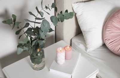 Photo of Vase with beautiful eucalyptus branches, book and candles on nightstand in bedroom