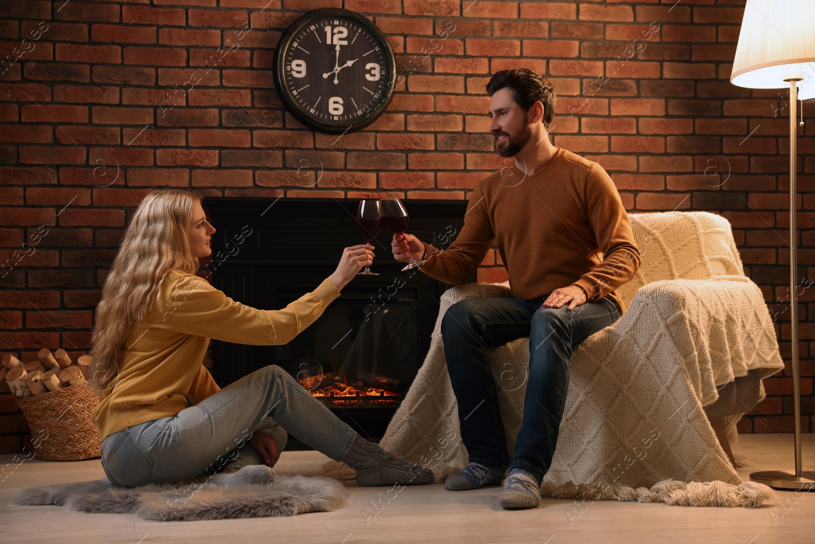 Photo of Lovely couple with glasses of wine spending time together near fireplace at home