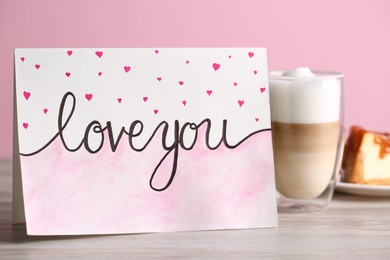 Photo of Card with phrase Love You and little drawn hearts, latte macchiato on white wooden table against pink background