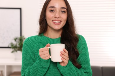 Photo of Beautiful young woman in stylish warm sweater holding cup of drink at home