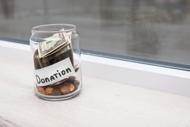 Photo of Glass jar with money and label DONATION on window sill. Space for text