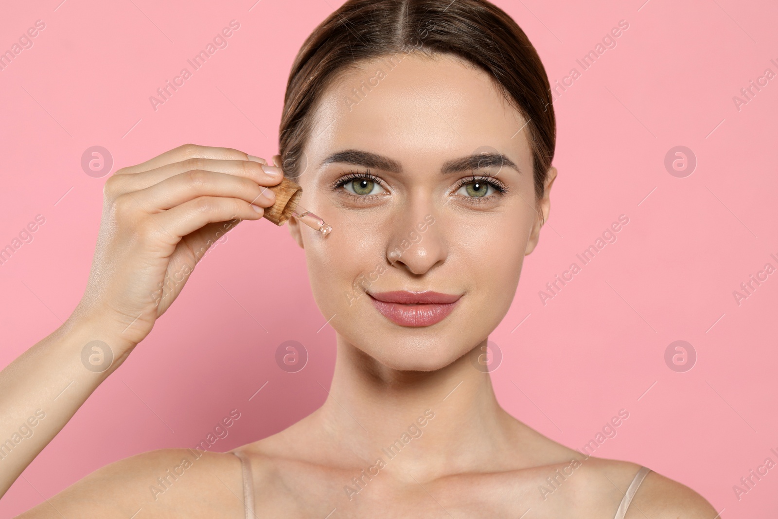 Photo of Young woman applying essential oil onto face on pink background