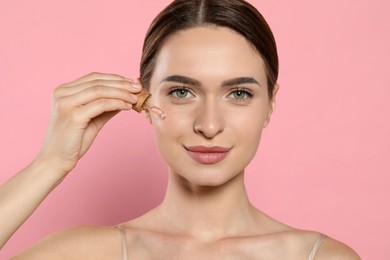 Photo of Young woman applying essential oil onto face on pink background