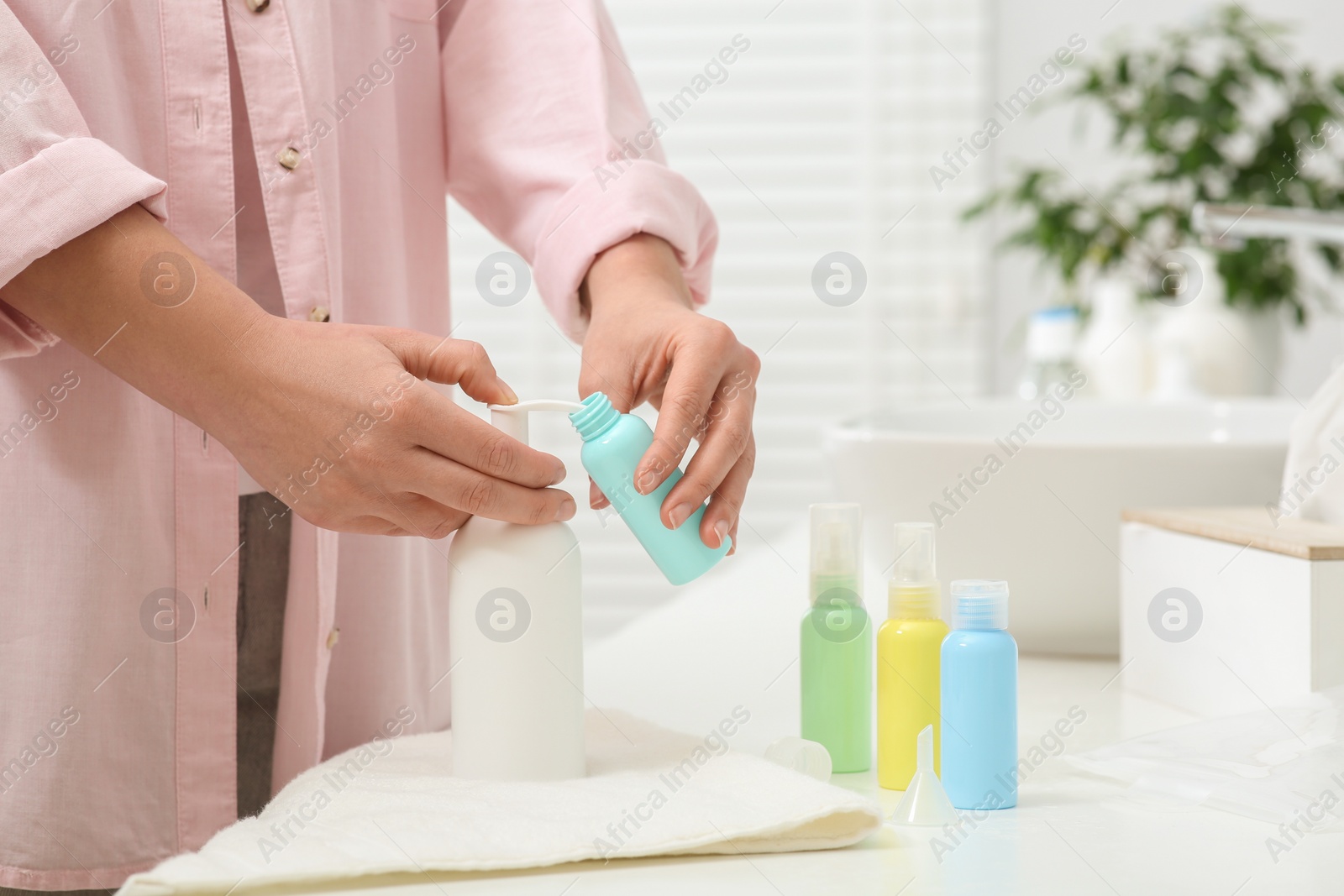 Photo of Woman pouring cosmetic product into plastic bottle at white countertop in bathroom, closeup. Bath accessories