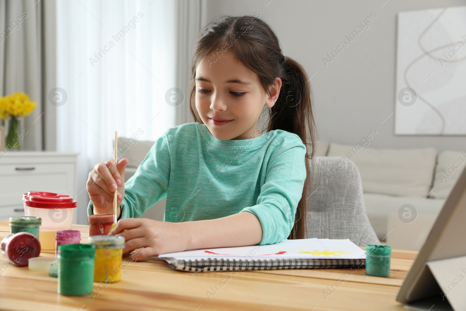 Photo of Little girl drawing on paper with paints at online lesson indoors. Distance learning