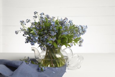 Photo of Bouquet of beautiful forget-me-not flowers in glass teapot and blue cloth on white table
