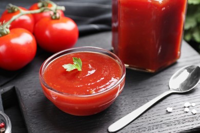 Photo of Delicious ketchup, spoon and salt on black wooden board, closeup. Tomato sauce