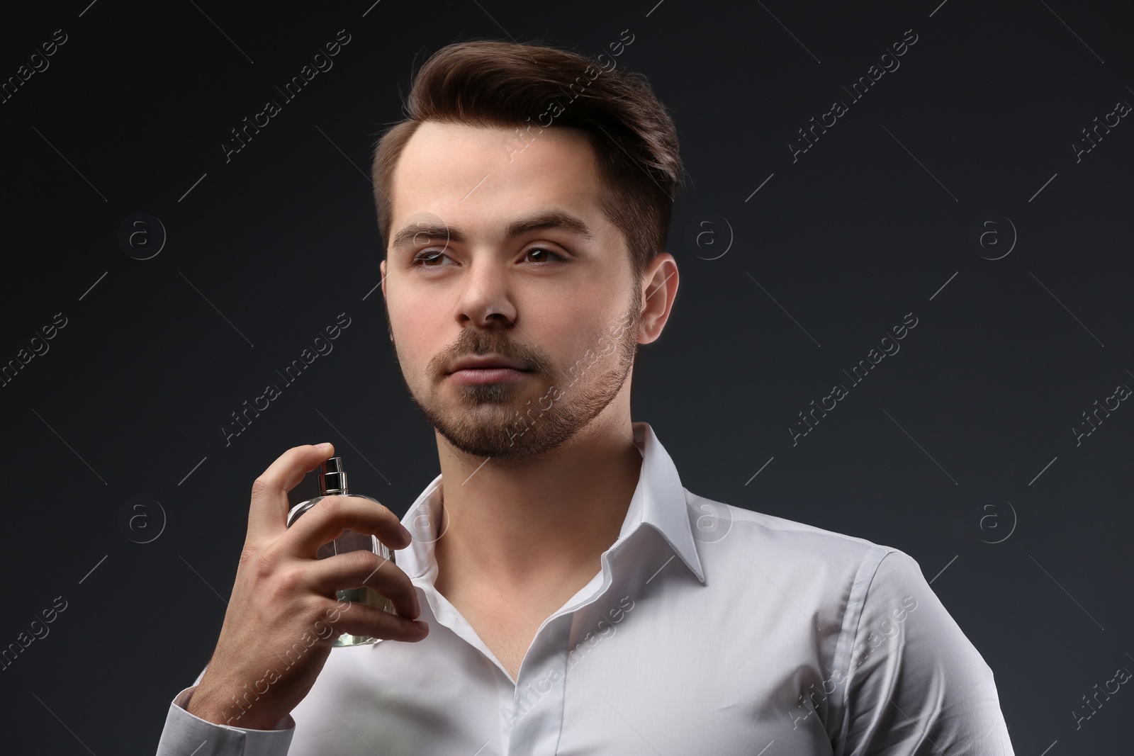 Photo of Handsome man in shirt using perfume on dark background